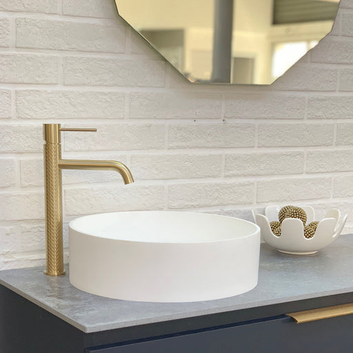 A Greens Textura Tower Basin Mixer in Brass standing by a white above counter basin